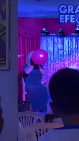 A person with blonde hair and a beard, wearing a dark dress, sits on a chair on a stage, blowing up a pink balloon. A man stands to their right, holding a microphone and gesturing. In the background, a blue tarp serves as a backdrop, with red curtains and a banner that reads "GRAN EFE SHOW". The stage is lit with pink and blue lights. In the foreground, two children are visible, facing away from the camera, watching the performance.