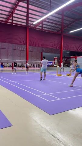 This video captures a mixed doubles badminton match played indoors. Four individuals are on the purple court, divided by a net. On one side, a man in a white t-shirt and blue shorts faces the net, while a woman in a blue sports bra and shorts is positioned further back, appearing to have just hit the shuttlecock. On the other side, a woman in a white and black athletic outfit stands closer to the net, and a man in a white t-shirt and black shorts is behind her.

The indoor setting features a high ceiling with exposed red support beams and translucent roofing panels allowing diffuse light. Large maroon drapes hang along the sides of the venue. The court is marked with white lines indicating boundaries. In the background, other badminton courts are visible, with more players and spectators. A banner hangs in the distance with the words "VICTOR CUP." The lighting suggests it is daytime, and the indoor environment implies it could be any time of day and unaffected by weather. The participants' postures and movements suggest an active and engaged match. The overall impression is one of a competitive sporting event within a dedicated indoor facility.