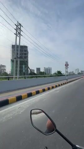 The video begins by showing a construction site with a tall building under construction, a road with yellow and black markings, and a speed limit sign. The camera then pans to the right, showing the road continuing with traffic cones and a traffic light. The video continues to show the road with decorative poles on the side and a clear sky with some clouds. A vehicle's side mirror is visible in the lower right corner, indicating the video is likely filmed from a motorcycle or scooter.