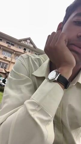 The video begins with a close-up of a person's face, partially obscured by their hand as they rest their chin on it. The person is wearing a light-colored collared shirt and a watch on their left wrist. In the background, a multi-story hotel with the word "IMPERIAL" visible on its facade is seen. The camera then pans to the left, revealing a well-maintained lawn and several dark-colored cars parked in front of the hotel. The scene shifts to a wider view of the hotel and its surroundings, showing more greenery, including manicured bushes and trees. The video continues to pan and zoom out, offering glimpses of more buildings and trees in the distance. The final frames show a row of tall pine trees and a single gold-colored chair placed on the grass. The sky is overcast, suggesting cloudy weather.