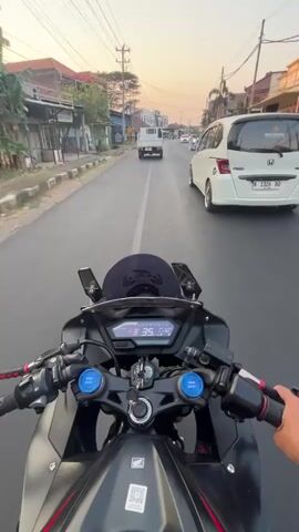 This is a first-person point-of-view video shot from a motorcycle. The motorcycle is black with red accents. The rider's hands are visible on the handlebars, and the digital speedometer displays a speed that increases from 35 to 39. The motorcycle is traveling on a paved road in what appears to be a semi-urban or suburban area. On the left side of the road are buildings and trees. On the right side of the road, there are other vehicles, including a white van, a white car, and a scooter with two people on it. The sky in the background is a pale orange, indicating it is either sunrise or sunset. The overall atmosphere suggests a casual ride through a town.