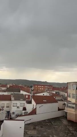 The video shows a cityscape under a grey, overcast sky. Several buildings with red tile roofs and light-colored facades dominate the scene. A few taller apartment buildings are also visible. In the distance, a line of hills can be seen. The weather appears stormy with a faint sound of thunder. The camera is static, offering a stationary view of the scene.