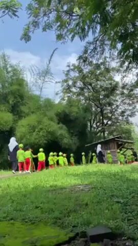 A group of about 20 children wearing matching green shirts, red pants, and green hats, hold hands and walk on a grassy path through a verdant outdoor area. The children are led by two adults dressed in black abayas and white hijabs. The landscape features bamboo, lush green trees, and mossy patches on the ground. The sky is bright with scattered clouds. There is a wooden structure with a roof visible in the background.
