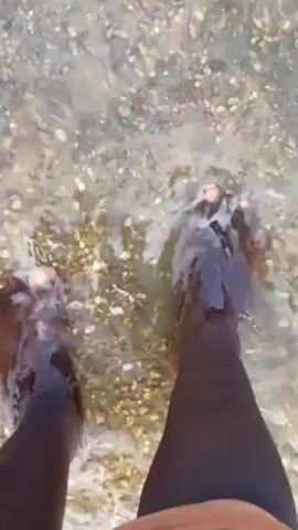 The video shows a first-person perspective of a person standing in shallow, clear water on a beach. The person's dark-skinned legs and feet are visible as the water gently laps around them. The seabed is covered in small pebbles and shells, visible through the clear water. The water is bright due to sunlight reflecting off the surface.