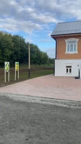 The video shows a traditional wooden house with a white foundation and a gray metal roof. The house has several windows, some with decorative white trim. In front of the house is a paved courtyard made of pinkish-gray pavers. To the left of the house, there is a wooden pole with a bell attached to it and a sign with some artwork. Behind the house, there are trees and a clear sky with some clouds. To the right of the house, there is a rustic wooden gate. The ground in front of the paved area is covered with gravel. The video pans from the left to the right, showcasing the house and its surroundings.