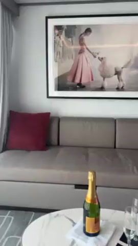 The video shows the interior of a cruise ship stateroom with a view of the sea and a building in the distance. The room has a sofa with a red pillow, a small white table, and a large framed photograph on the wall. White sheer curtains hang next to the sliding glass door, which leads to a balcony. The balcony has two chairs and a small table. The water appears turquoise, and the sky is partly cloudy.