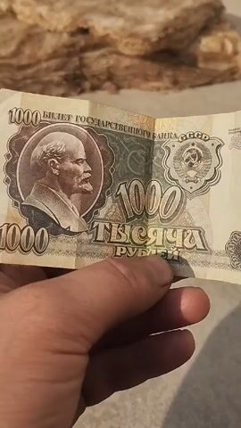 A hand holds a 1000 Ruble banknote featuring a portrait of Lenin. The bill has text in Russian and displays the Soviet Union's coat of arms. The background is blurry, showing a beige, fiber-like material. The scene is brightly lit by daylight.