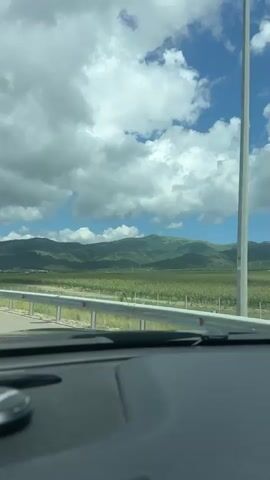 A view from inside a vehicle shows a landscape with a road, guardrail, green fields, and rolling hills under a blue sky with white clouds. The interior of the car is visible in the foreground, with a dashboard and part of a seat. There are no people visible. The video appears to be shot from a moving vehicle, with the landscape changing slightly as the car progresses.