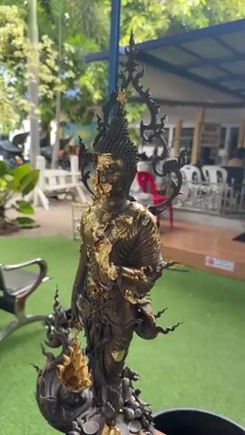 The video shows a close-up, slow pan of a bronze statue of a standing Buddha adorned with gold leaf. The statue rests on a decorative base resembling a mythical creature, possibly a Naga, with coins scattered around its feet. The statue is predominantly dark bronze with gold accents, particularly on the face and robes. The background is blurred, suggesting an outdoor setting, with hints of greenery, a building, and other objects that are out of focus. The lighting is natural and somewhat bright.