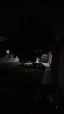 The video shows a dark outdoor scene at night. A building with some windows is visible on the left. There are trees and possibly some vehicles in the middle and right of the frame. The lighting is dim, with a few scattered light sources. The footage is shaky and somewhat blurry, suggesting it was recorded while moving.