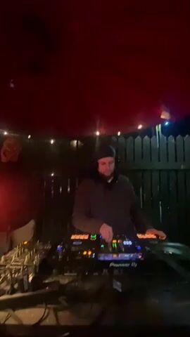 A DJ wearing a black beanie and headphones operates a Pioneer DJ console outdoors.  He manipulates the controls, blending music with a deep bassline.  Behind him, a person in a light-colored shirt stands watching, partially obscured by shadows.  The scene is dimly lit, with a red cloth overhead and string lights along a dark wooden fence in the background. The area around the DJ console is cluttered with cables.