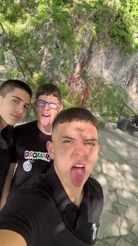 The video starts with three young men making faces in a selfie. The background shows stone steps, a bench with people sitting on it, trees, and a rocky cliff with some greenery. The video transitions to show a young man walking along a stone walkway, with a railing on his right, and a rocky cliff on the left. He turns the camera to show a large waterfall cascading down the cliffside. He smiles at the camera. The surroundings are lush with greenery, with trees framing the waterfall. The sky is visible through the treetops.