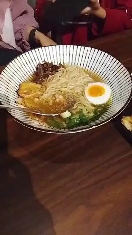 The video shows a close-up view of a table with food. In the foreground, there is a bowl of ramen with noodles, broth, a boiled egg cut in half, and some greens. To the right of the ramen bowl is a small dish of dark sauce and a plate of fried enoki mushrooms. In the background, two people are partially visible, one wearing a red sweater and the other in a patterned garment. One of the individuals appears to be holding a phone. The table surface is a dark wood grain. The scene appears to be indoors, likely a restaurant. The camera angle is from a slightly elevated perspective looking down at the table.