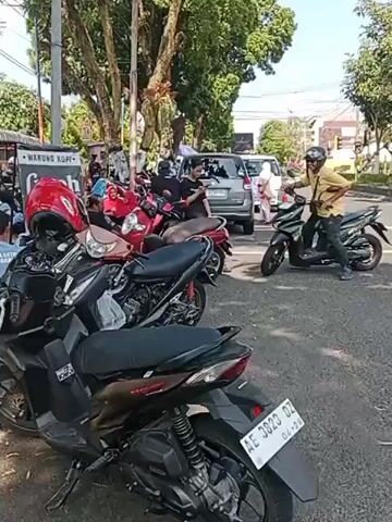 A man wearing a yellow shirt and a helmet sits on a black motorcycle. He is looking to his right. Several other motorcycles are parked in front of him. In the background, there are trees and buildings. There are also orange traffic cones on the side of the road. The video is taken from a low angle, looking up at the man and the motorcycles.
