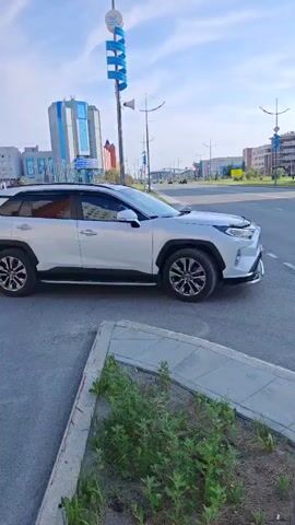 The media shows a white Toyota RAV4 driving on a paved road in a modern urban environment. The car is the primary object of focus, seen from the side and then the rear as it moves away from the camera. The setting features several contemporary buildings with geometric architectural designs, suggesting a relatively new development area. A blue and white sign with spiral accents stands to the left of the road, and a lamppost is visible in the foreground. The sky is blue with scattered clouds, indicating a clear day, likely in the late morning or afternoon due to the bright lighting. The overall mood is neutral, capturing a mundane moment of vehicular traffic. License plate information visible on the rear of the car reads "A 747 EA 177 RUS". A speed limit sign with "40" is also visible on the right side of the road. The presence of green grass and some weeds along the roadside suggests an outdoor setting, possibly a suburban or commercial district.