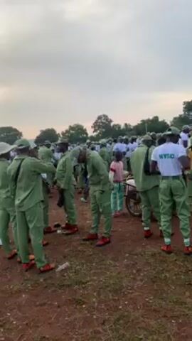The video shows a group of people dressed in green and white NYSC (National Youth Service Corps) uniforms standing in a field. Some are wearing caps, while others are not. The uniforms have "NYSC" printed on the back. There are also several children in the area, some dressed in civilian clothing. One child is pushing a wheelbarrow. The people in uniform appear to be milling around and interacting with each other. The field is mostly brown dirt, with some patches of grass. In the background, there is a building and some trees, and the sky is overcast.