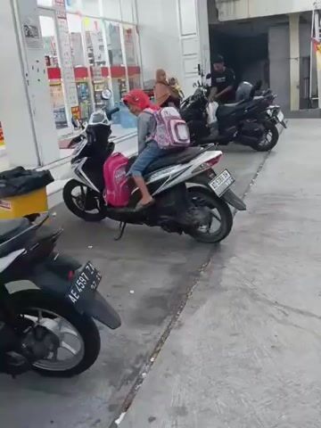The video shows an outdoor scene with several motorcycles parked on a concrete surface in front of a building with shelves stocked with goods. In the foreground, a black and white motorcycle is parked. To the right, a white and black motorcycle is parked, with a person wearing a pink hijab and a backpack sitting on the seat. The person is looking back and smiling. To the right of this motorcycle, another black motorcycle is parked, with a man wearing a black shirt standing beside it, holding a plastic bag. In the background, a woman wearing a hijab and a child are standing near the building. The building appears to be a store or a convenience store, indicated by the shelves of products visible through the glass facade. The overall setting suggests a public area, possibly a parking lot or an area outside a commercial establishment.