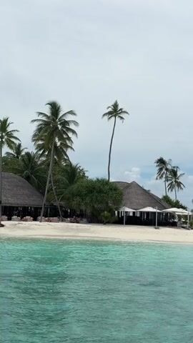The video shows a tropical beach scene. In the foreground, there is turquoise water. In the middle ground, there is a white sand beach with palm trees. There are thatched-roof buildings, umbrellas, lounge chairs, and a hanging rattan chair visible on the beach. Several people are seated on lounge chairs or walking on the beach. In the background, there is a cloudy sky. The camera appears to be moving slightly to the right.