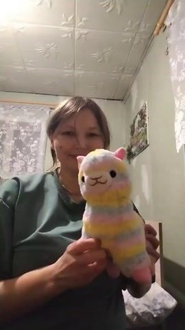A woman is holding a stuffed animal llama up and down in front of a camera. The llama is multi-colored, with stripes of yellow, grey, pink, and white. It has pink ears. The woman is wearing a green shirt and has brown hair. She is smiling. The background includes a patterned ceiling, a window with lace curtains, and a picture on the wall. The lighting is somewhat dim.