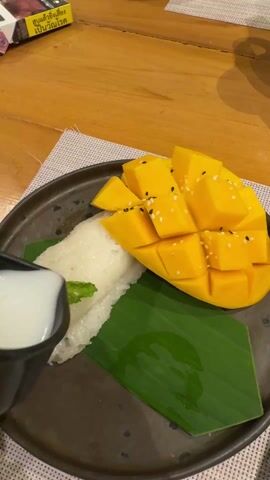 Here's a detailed description of the video:

The video shows a plate of mango sticky rice, a popular Southeast Asian dessert. A hand is pouring coconut milk from a small black cup over the mango and sticky rice. The mango is cut into a grid pattern and sprinkled with black sesame seeds. The sticky rice is garnished with a few mint leaves. The dessert sits on a green banana leaf, which rests on a dark brown plate. The plate is placed on a wooden table covered with a woven placemat. In the background, a pack of cigarettes is partially visible. The lighting appears to be from an indoor setting.