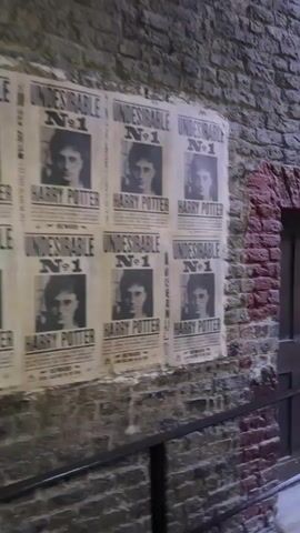 The video shows a cobblestone alleyway with brick walls on either side.  Several wanted posters featuring the face of a young man are affixed to the left wall.  A wooden door is set into the right wall.  Further down the alley, the scene opens up to a street lined with shops.  A gas lamp illuminates the area.  Several people are walking through the street, some appearing to be tourists.  One person in the foreground is wearing a white t-shirt and dark shorts. Another person is wearing a red dress. The buildings have upper floors with windows. The overall environment suggests a themed attraction or a movie set.