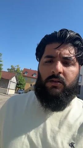 A man with a beard is recording a video of himself and a woman walking down a cobblestone street on a sunny day. The man is wearing a light yellow t-shirt. The woman is wearing a white t-shirt and blue jeans. She is holding a blue bag. They are walking towards the camera. The street is lined with trees and houses. The sky is blue.