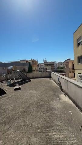 A vertical, eye-level shot shows a rooftop scene under a clear blue sky with the sun prominently visible. The rooftop itself is made of concrete and appears weathered, with stains and a visible drain in the foreground. Two solar water heaters are positioned on the rooftop; one is a darker, sloped panel with a circular component at the top, while the other is a similar panel angled more steeply, also with a circular component. Several metal rods protrude from the concrete near the solar heaters. In the background, buildings of varying heights and architectural styles are visible, suggesting an urban or suburban environment. Some buildings have balconies, air conditioning units, and solar panels on their roofs. The overall impression is one of a sunny day in a populated area, with practical rooftop installations.