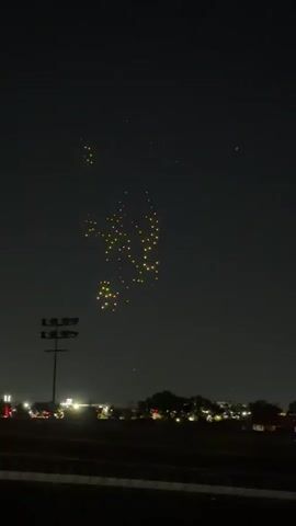 A drone show displays a lion rampant crest within a shield shape. The crest is rendered in red and yellow lights, while the shield outline is primarily red. The drones are arranged in a grid formation against a dark night sky. Below the drone display, the lights of a distant town or city are visible on the horizon, along with a single streetlamp or light pole in the foreground. The image captures a dynamic moment of the drone performance as the shield shape solidifies.
