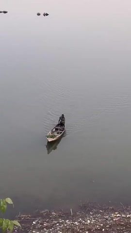 A small wooden boat with two people inside is being rowed across a wide body of water. The water is calm with gentle ripples. In the background, there are hills and sparse vegetation. The foreground shows the water's edge with debris.