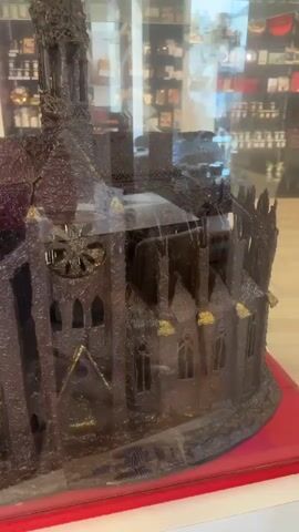 A close-up shot focuses on a chocolate sculpture of a cathedral, displayed behind glass. The sculpture is dark brown and intricately detailed, with spires, arches, and what appear to be stained-glass windows depicted in the chocolate. Some gold accents are visible on the sculpture. The background is blurred, showing shelves stocked with various items, possibly in a shop. The camera slowly pans around the sculpture.