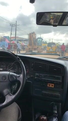 This media file captures a street scene from inside a car in Aketu-Oja, Nigeria. The camera is positioned behind the steering wheel of a Volkswagen, looking out through the windshield.

The primary subjects are the people and vehicles present in the bustling outdoor environment. A large yellow excavator is a prominent object in the middle of the road, suggesting some form of construction or road work. Surrounding it, there is a lively market scene with numerous stalls covered by colorful umbrellas, and many people walking around, some wearing bright clothing. Several other vehicles are visible, including a yellow motorized tricycle (Keke Napep) and other cars.

The activity appears to be a typical day in a busy Nigerian marketplace, with people going about their business amidst traffic and construction. The time of day appears to be daytime, likely late morning or early afternoon, given the lighting conditions. The sky is overcast with thick clouds, indicating potentially dull or rainy weather.

Notable details include the interior of the car, showing the dashboard, steering wheel, and radio. The side mirror reflects individuals, possibly passengers or bystanders. The presence of power lines and poles suggests an urban or semi-urban setting. The overall atmosphere conveyed is one of lively activity and everyday life in a community.

There is some visible text on a sign in the background, but it is difficult to read clearly. The overall scene depicts a dynamic and somewhat chaotic yet ordinary moment in a Nigerian town.