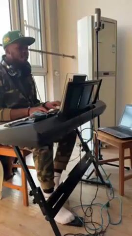 A man wearing a camouflage jacket, pants, and a green baseball cap sits on a stool, playing a black electric keyboard. He has headphones around his neck. The keyboard is on a black metal stand. A music stand is attached to the keyboard, with papers on it. A microphone on a stand is to the right of the keyboard. A laptop is on a small wooden table to the right of the keyboard. Several cords are visible on the floor. A large air conditioning unit is behind the microphone. Light comes in through a large window to the left. The floor appears to be wood.