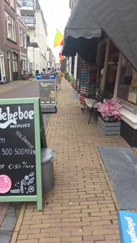 The video shows a view from street level looking down a narrow cobblestone street. On the left is a sidewalk with a green A-frame chalkboard sign in the foreground. The sign reads "Kiekeboe snoepgoed" and lists "Ruim 500 soorten" (Over 500 varieties) and prices starting from €1.50 per 100 grams, with illustrations of a lollipop and candy canes. A trash bin is next to the sign. Further down the street on the left are buildings with windows and flags hanging from them. On the right side of the street is another sidewalk in front of shops. A black metal bench with a white cushion is visible on the right sidewalk, next to a grey wooden planter filled with pink artificial flowers. There are racks of colorful items displayed outside the shops. In the distance, a few people are walking and cycling, and there is a small cart with colorful items on the street. The buildings are old-fashioned, with multiple stories. The sky appears overcast.