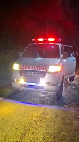 A white Suzuki ambulance is parked on the side of a dirt road at night. The ambulance has its emergency lights activated, with red lights on the roof and blue and yellow lights on the front. The word "AMBULANCE" is written in red letters on the front of the vehicle. The side door of the ambulance is open, and a person wearing dark clothing is visible inside. The background is dark, with trees and foliage visible. The ground in front of the ambulance is lit by the vehicle's lights, casting yellow and blue glows.