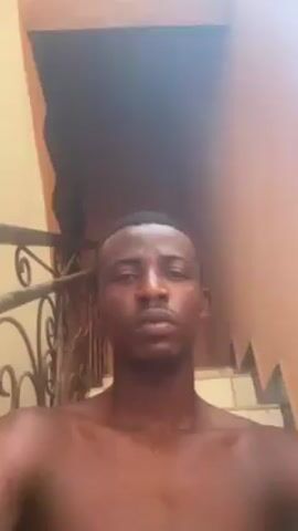 The video shows a close-up shot of a dark-skinned man with short, dark hair and a short beard. He is shirtless and looking directly at the camera. The background includes a light-colored wall, a metal railing with a decorative design, and stairs. The lighting is bright.