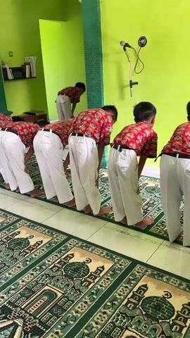 A group of male children, dressed in red and white school uniforms, are standing in a line on a green and white patterned rug. They are all facing the same direction and appear to be performing a religious ritual, specifically the bowing posture known as Ruku in Islamic prayer. A microphone is mounted on the wall behind them. The walls of the room are painted green. In the background, another child in the same uniform is standing, facing away from the camera. One child in the foreground is reaching out with their left hand towards the child in front of them. The overall scene suggests a communal prayer or religious lesson taking place in a mosque or prayer room.