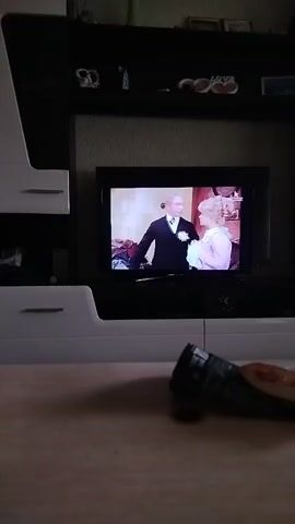 A television screen displays a scene from a period drama. A man in a dark suit and a woman in a light-colored dress stand close together. In another scene, a man in a light-colored suit is visible. The television is mounted on a wall unit with shelves above it. In the foreground, a blurred object resembling a wrapper or package lies on a table.