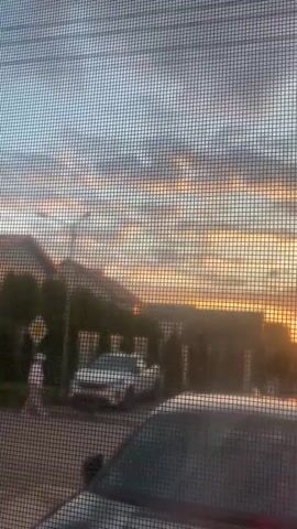 This is a video of a sunset viewed through a window screen. The sky is filled with clouds, some dark gray and others tinged with orange and yellow from the setting sun. In the foreground, the mesh of the window screen is clearly visible, creating a grid-like pattern over the scene. Behind the screen, there are houses, trees, and cars. One white car is parked in front of a house, and another car, possibly dark-colored, is closer to the viewer. A person in a white outfit is walking on a sidewalk. A yellow traffic sign is visible on the left side of the image. The overall impression is a suburban street scene at dusk.