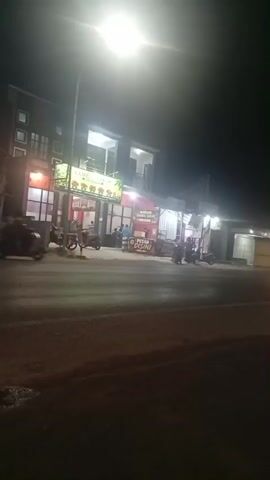 A nighttime scene unfolds along a roadside. In the foreground, a dark, wet asphalt road stretches across the frame, with patches of illuminated water reflecting the overhead streetlights. Beyond the road, a row of buildings lines the street. The closest building features a brightly lit storefront with red and white accents and a sign displaying colorful fruit imagery. Several motorcycles are parked in front of this establishment, and a few individuals are visible inside and around the entrance. Further down the street, more businesses appear, also illuminated by artificial light. A large truck, its headlights and taillights blurred by motion, drives past in the middle ground, temporarily obscuring the view of the buildings. The overall ambiance is that of a commercial area active during the evening hours, with the glow of lights and the presence of vehicles and pedestrians.