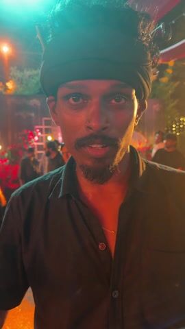 A man with dark skin, a beard, and a black wrap around his head is standing in front of a crowd of people. He is wearing a black button-down shirt and a gold chain. The background is decorated with lights and flowers. The man is talking and gesturing with his hands. He smiles.