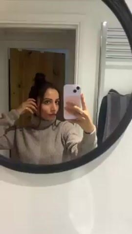 A woman with her hair in a bun is holding a phone in front of a bathroom mirror and is taking a selfie. She is wearing a taupe-colored turtleneck sweater and is standing in front of a white wall. Behind her is a doorway leading to another room with a wooden door. To the right of the mirror is a heated towel rack with a gray towel hanging on it. The bathroom has white walls and appears to be well-lit. The woman is looking at the camera.