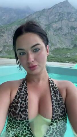 A woman with dark hair and blue eyes, wearing a leopard print one-piece swimsuit, poses for a selfie in a hot tub with mountains in the background. The water is a light blue, and the sky is overcast. There is a small, lit candle on the edge of the hot tub to the right.