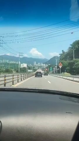 The video shows a first-person perspective from inside a car driving on a road. The car is moving forward on a highway or a wide road with multiple lanes. In front of the car, other vehicles are visible, including a black car and a white SUV. The road is bordered by a yellow and black guardrail on the left and a white barrier with yellow poles on the right. On the left side of the road, there are power lines and a line of trees. In the distance, mountains and buildings can be seen under a clear blue sky with some clouds. Traffic signs are visible along the road, including a sign with the number 46. The interior of the car is visible in the foreground, with the dashboard and windshield. The overall environment appears to be a suburban or semi-rural area during the daytime.