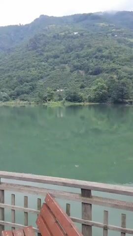 The video shows a serene lake surrounded by lush green mountains. A wooden deck with a railing overlooks the water, and a bench is placed on the deck. The water is calm and reflects the surrounding landscape. The sky is overcast, suggesting a pleasant day for outdoor activities. The overall atmosphere is peaceful and tranquil.
