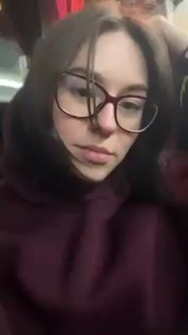 A young woman with shoulder-length dark hair and wearing maroon-framed glasses and a maroon hoodie sits in a bus seat. She looks directly at the camera, then down, and then to the side. The seat and window beside her are red and black. She appears to be indoors, likely traveling.