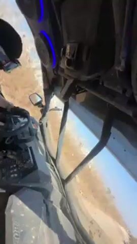 The video shows a first-person view from inside a Can-Am side-by-side vehicle driving on a dirt road. The driver is wearing gloves and appears to be maneuvering the vehicle, as evidenced by the visible steering wheel and gear selector. The environment consists of a dry, grassy field under a clear blue sky. The vehicle is equipped with a windshield and side mirrors. There is some apparent dust or dirt on the vehicle, suggesting it has been used off-road.