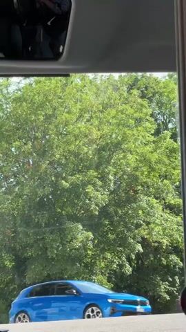 The video shows a view from inside a vehicle, likely a bus, looking out the window. The first few seconds show lush green trees and a bright sky. Then, a blue car is visible passing by. The scene shifts to show a white building with two windows and a tiled roof. A street sign labeled "Route de Bell" is also visible. Finally, the view shows a wooden house under construction with a red tiled roof and several cars on a road in front of it. The overall environment appears to be a town or village with a mix of residential and possibly commercial buildings, surrounded by trees and greenery. The lighting suggests it is daytime.