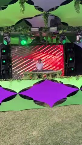The video shows a DJ in a white shirt and glasses standing behind a mixing board on a stage with a large video screen behind him. The screen displays red and white visuals. The stage is decorated with green plants, and the structure above the stage is made of green and black fabric panels. Below the stage, there are additional panels of green and purple fabric. The stage is set up on a grassy field. There are speakers visible on either side of the DJ. The overall impression is that this is a music performance at an outdoor event or festival.