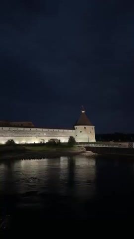 The video shows a nighttime view of a historical stone fortress situated on the bank of a river. The fortress is illuminated by artificial lights, casting a warm glow on its walls and a prominent tower. The river in the foreground reflects the lights and the dark sky, creating a calm and serene atmosphere. The sky is dark with some faint cloud formations visible. The scene appears to be a static shot, with no visible movement within the frame, other than the subtle ripples on the water's surface.