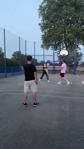Four people are playing volleyball on an outdoor court. The court is paved and has a basketball hoop at one end. A blue fence surrounds the court. The sky is a pale blue, suggesting it is either early morning or late evening. In the foreground, a person wearing a black t-shirt and grey shorts stands with their back to the camera, facing the net. A ball is in the air. To the left, a person in an orange t-shirt and white shorts is about to hit the ball. In the middle ground, a person wearing a black t-shirt and grey pants stands facing the net. To the right, a person in a pink t-shirt and black shorts is in the process of hitting the ball over the net. Another person in a white t-shirt is visible in the far right of the frame.